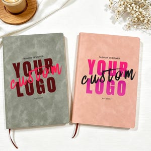 May include: Two rectangular notebooks with textured covers, one in sage green and one in soft pink. Each features the text FASHION DESIGNER, YOUR CUSTOM LOGO, and EST 2035 printed on the front. The green notebook has dark red text, while the pink notebook has bright pink and black text. Both include a thin ribbon bookmark.