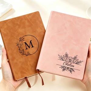 Op de afbeelding: Twee rechthoekige notitieboeken met gladde covers. Het linker notitieboek is lichtbruin en heeft een zwart cirkelvormig bloemmotief met de letter M. Het rechter notitieboek is lichtroze en heeft een zwarte bloemafbeelding met de naam Chelsea.