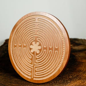 May include: A round, wooden labyrinth with a carved design. The labyrinth is made of light brown wood and features a detailed pattern of concentric paths and a central four-leaf clover design. The wood has a natural grain and is displayed on a dark wood surface.