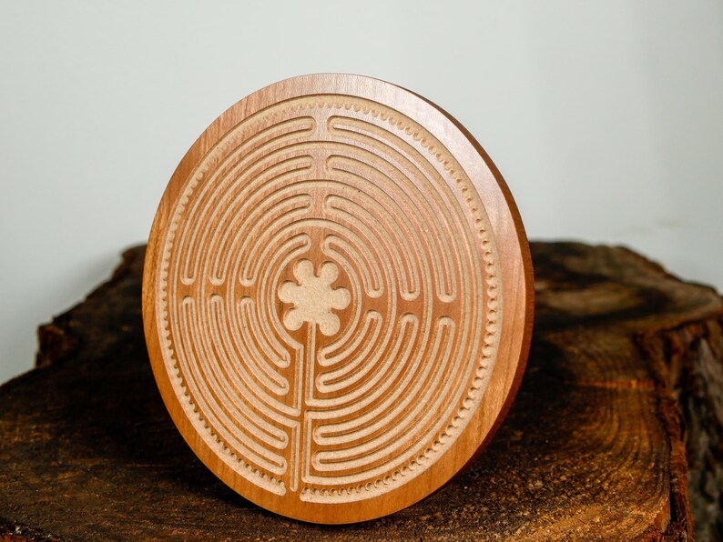 May include: A round, wooden labyrinth with a carved design. The labyrinth is made of light brown wood and features a detailed pattern of concentric paths and a central four-leaf clover design. The wood has a natural grain and is displayed on a dark wood surface.