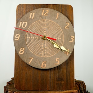 May include: A wooden desk clock with a round face and a rectangular base. The clock face is light brown with etched numbers and a maze-like design in the center. The clock hands are gold, and the second hand is red.