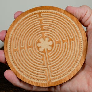 May include: A round, wooden coaster with a carved labyrinth design. The coaster is light brown with a flower-like shape at the center of the maze. The coaster is held in a person's hand.