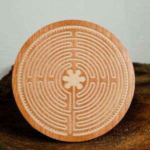 May include: A round wooden labyrinth with a carved design. The labyrinth features concentric circles and a central floral shape. The wood has a light brown color and is set on a dark brown, textured surface.