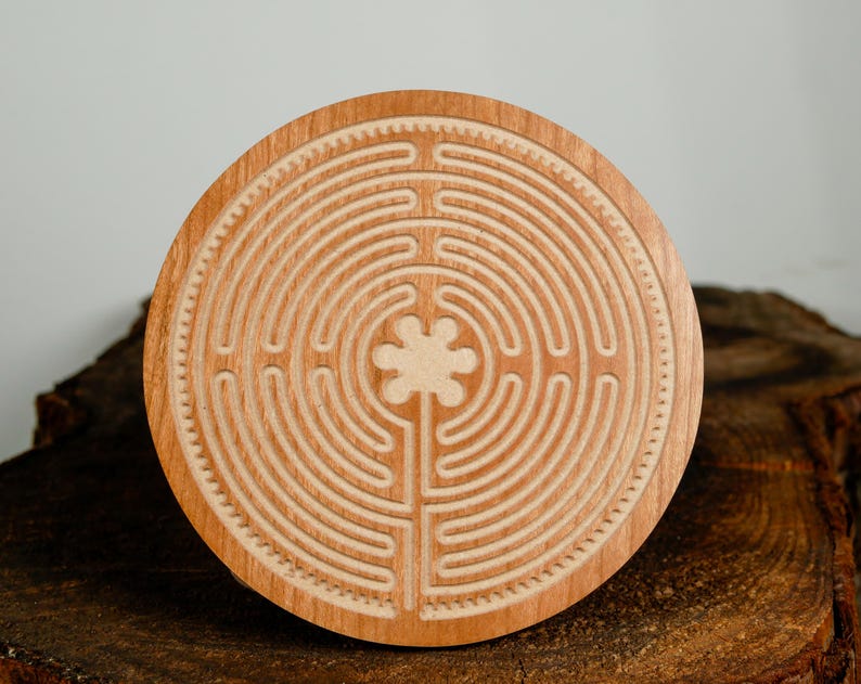 May include: A round wooden labyrinth with a carved design. The labyrinth features concentric circles and a central floral shape. The wood has a light brown color and is set on a dark brown, textured surface.