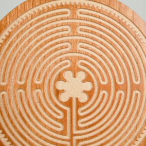 May include: A circular wooden labyrinth with a carved path design. The labyrinth is made of light brown wood and features a series of concentric circles and a central floral shape. The outer edge has a decorative gear-like pattern.