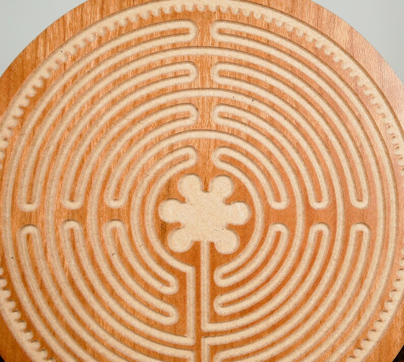 May include: A circular wooden labyrinth with a carved path design. The labyrinth is made of light brown wood and features a series of concentric circles and a central floral shape. The outer edge has a decorative gear-like pattern.