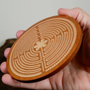 May include: A round, wooden coaster with a carved labyrinth design. The coaster is made of light brown wood and features a detailed pattern of concentric circles and a central floral element. The coaster is held in a hand.