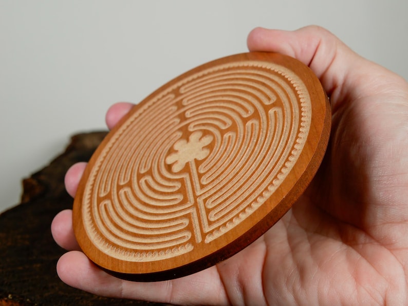 May include: A round, wooden coaster with a carved labyrinth design. The coaster is made of light brown wood and features a detailed pattern of concentric circles and a central floral element. The coaster is held in a hand.