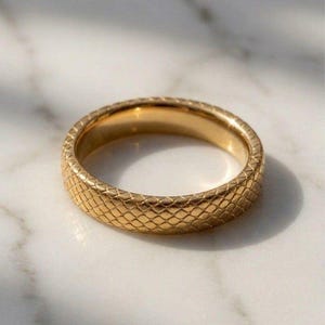 May include: A gold-toned band ring featuring a textured, repeating diamond-shaped pattern across its outer surface. The ring has a polished finish and a consistent width, showcasing a metallic sheen that reflects light across its geometric design.