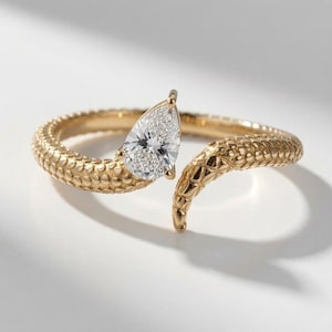 May include: A gold ring featuring a textured, scale-like band that wraps around the finger in an open design. One end of the band is topped with a single, clear, pear-shaped gemstone that sits prominently at the center of the ring's opening.