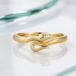May include: A gold ring shaped like a coiled snake with a textured scale pattern covering the band. The snake head features two small, clear, round inset stones representing eyes, and the tail curves inward to complete the circular band design.