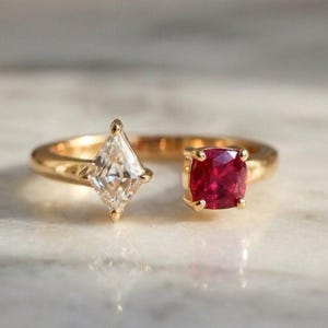 May include: A gold open-style ring featuring two distinct gemstones. On one side, a clear, kite-shaped diamond is set in a prong setting. On the opposite side, a vibrant, square-cut red ruby is secured in a matching gold prong setting.