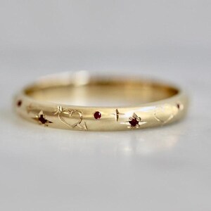 May include: A gold band ring featuring engraved heart and star motifs, accented with small, round, deep red gemstones set into the metal surface. The design includes a mix of etched heart outlines and starburst shapes across the band.