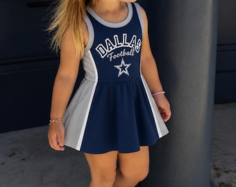 Può includere: Un abito senza maniche blu navy e grigio con la scritta Dallas Football e una grafica a stella sul petto. L'abito presenta una gonna svasata con pannelli laterali grigi a contrasto e bordini bianchi lungo lo scollo e i giromanica.