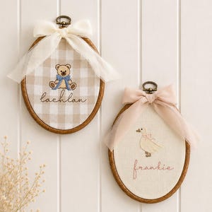 May include: Two oval wooden embroidery hoops featuring fabric inserts. The left hoop has beige gingham fabric with an embroidered teddy bear and the name lachlan, topped with a white ribbon. The right hoop has cream fabric with an embroidered goose and the name frankie, topped with a pale pink ribbon.