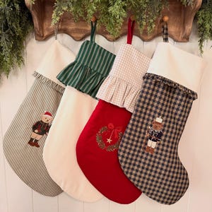 May include: Four Christmas stockings hanging in a row. The first is grey striped with a teddy bear, the second is solid cream with a green striped ruffle, the third is red with a wreath and gingham ruffle, and the fourth is navy plaid with a bear.