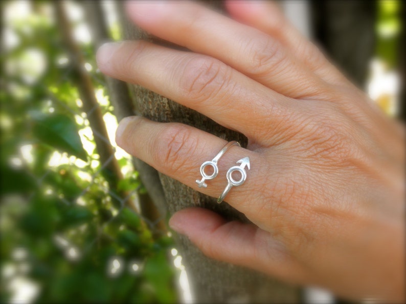 May include: Two silver rings with a simple design. One ring features a circle with an arrow pointing to the right. The other ring features a circle with a symbol that resembles the male gender symbol.