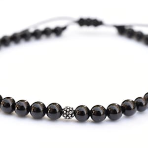 Puede incluir: Un collar de cuentas negras con una cuenta plateada en el centro. El collar est&aacute; hecho de cuentas peque&ntilde;as y redondas y est&aacute; atado con un cord&oacute;n negro.
