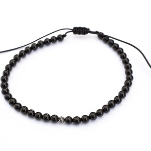 Puede incluir: Un brazalete de cuentas negras con una cuenta plateada en el centro. El brazalete es ajustable con un nudo deslizante.