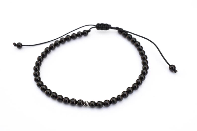 Puede incluir: Un brazalete de cuentas negras con una cuenta plateada en el centro. El brazalete es ajustable con un nudo deslizante.