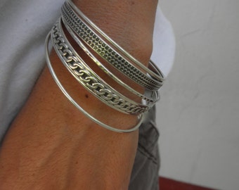 Puede incluir: Un conjunto de cinco pulseras de plata con diferentes texturas y patrones. Las pulseras están apiladas en la muñeca de una persona.