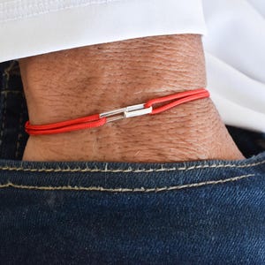 May include: A red cord bracelet with a silver clasp. The bracelet is worn on a wrist, with a white shirt and blue jeans in the background. The bracelet is a simple, minimalist design.