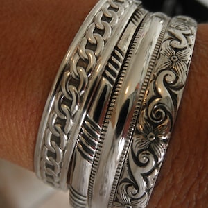 May include: A set of four silver bracelets with intricate designs. The bracelets are stacked on a person's wrist. The top bracelet has a chain-like design, the second has a geometric pattern, the third is a plain band, and the bottom bracelet has a floral design.