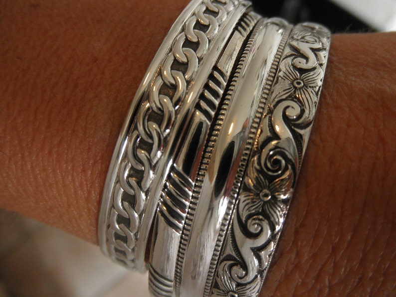 May include: A set of four silver bracelets with intricate designs. The bracelets are stacked on a person's wrist. The top bracelet has a chain-like design, the second has a geometric pattern, the third is a plain band, and the bottom bracelet has a floral design.
