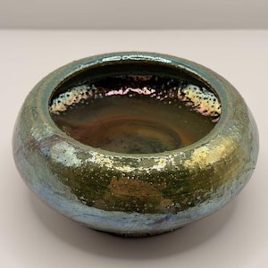 May include: A round ceramic bowl with a wide opening and a low profile. The exterior features a textured, iridescent glaze with blended shades of metallic green, blue, and gold. The interior is dark and reflective with a smooth, glossy finish.