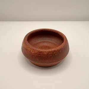 May include: A round, low-profile ceramic bowl with a textured, mottled surface in shades of deep reddish-brown and burnt orange. The interior is smooth and glossy, while the exterior features a rough, cratered glaze with a distinct horizontal ridge.