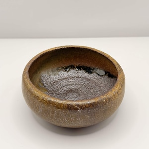 May include: A round ceramic bowl with a textured, mottled brown and tan exterior. The interior features a dark, glossy, and uneven glaze that pools at the bottom, contrasting with the rough, earthy appearance of the outer rim and sides.