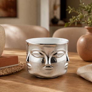 May include: A cylindrical ceramic planter with a reflective silver finish. The exterior surface features molded, stylized human faces with closed eyes, noses, and lips embossed around the entire circumference. The interior is smooth and white.
