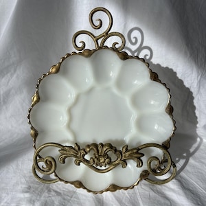 May include: A white milk glass egg serving platter with a scalloped edge, held by an ornate antique brass metal stand. The stand features intricate scrollwork and a central fleur-de-lis motif, providing a decorative frame for the twelve-well plate.
