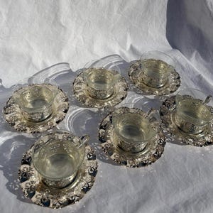 May include: A set of five clear glass teacups with intricate silver-toned metal holders and matching silver-toned saucers. Each cup features a delicate floral cutout pattern on the metal base and saucer rim, creating a cohesive and reflective appearance.