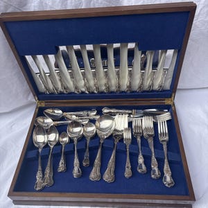 May include: A silver-toned cutlery set arranged in a wooden storage case with a blue velvet interior. The set includes various spoons, forks, and knives with ornate handles. The case features a hinged lid with a mirror panel and slots.