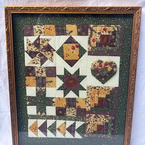 May include: A framed fabric wall hanging featuring a patchwork quilt design with various geometric blocks including stars, pinwheels, and a heart. The fabric patterns incorporate shades of mustard yellow, deep brown, cream, and forest green.