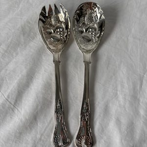 May include: A pair of silver-toned metal salad servers featuring a polished finish. The set includes one slotted spoon and one solid spoon, both decorated with an embossed pineapple and leaf motif on the bowl and a fluted design on the handle.