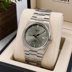 May include: A Tissot PRX Powermatic 80 wristwatch with a brushed stainless steel case and integrated bracelet. The watch features a textured green waffle dial, silver-tone hands and indices, and a date window at the three o'clock position.