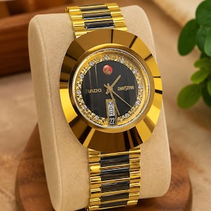 Op de afbeelding: Een Rado DiaStar polshorloge met een zwarte wijzerplaat, goudkleurige wijzers, een dag-datumvenster met MAR 22 en een goudkleurige ring bezet met kleine heldere steentjes. De schakelarmband heeft afwisselend gouden en zwarte schakels.