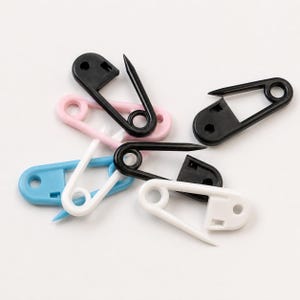 Op de afbeelding: Een verzameling plastic veiligheidsspelden in diverse kleuren, waaronder zwart, wit, lichtblauw en lichtroze. Elke speld heeft een puntig uiteinde en een sluitmechanisme, gerangschikt in een hoopje tegen een neutrale achtergrond.