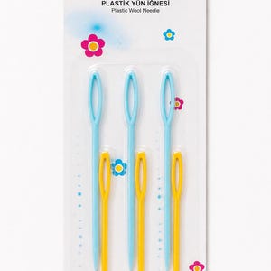 Op de afbeelding: Een verpakking met zes plastic wolnaalden, bestaande uit drie lichtblauwe en drie gele naalden met grote ogen. De witte verpakking bevat de tekst MC PLASTIK YÜN İĞNESI Plastic Wool Needle en 6 Adet, versierd met kleine kleurrijke bloemiconen.