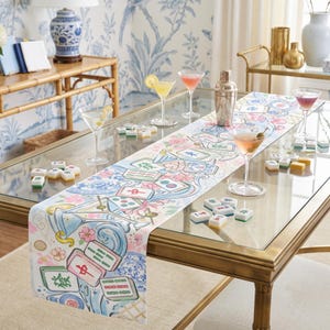 May include: A white table runner featuring a colorful illustration of mahjong tiles, martini glasses, and pink cherry blossoms. The design includes blue swirling water patterns and scattered mahjong tiles across the fabric surface.