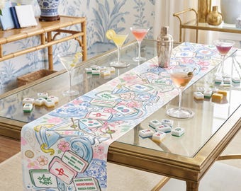 May include: A white table runner featuring a colorful illustration of mahjong tiles, martini glasses, and pink cherry blossoms. The design includes blue swirling water patterns and scattered mahjong tiles across the fabric surface.