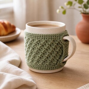 Puede incluir: Una taza de cerámica con acabado moteado en color blanco roto, llena de una bebida de color marrón claro. La taza está envuelta en una funda de ganchillo texturizada de color verde salvia con un patrón de punto de concha que rodea el cuerpo y el asa.