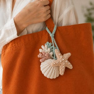 May include: A burnt orange tote bag featuring a hanging crochet accessory. The accessory consists of a light blue cord attached to a cluster of crocheted sea-themed items, including a white seashell, a beige starfish, and small pink and white coral pieces.