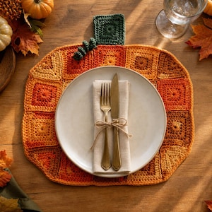 Puede incluir: Un mantel individual con forma de calabaza hecho de cuadrados de ganchillo en tonos naranja, amarillo y marrón, con un tallo de ganchillo verde oscuro. Un plato de cerámica blanca descansa sobre el mantel con una servilleta beige, un tenedor y un cuchillo dorados.