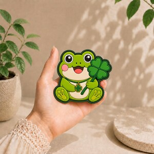 Può includere: Una toppa a forma di rana verde con pancia color crema, guance rosa e un ampio sorriso. La rana tiene in mano un grande quadrifoglio e indossa una collana con un piccolo ciondolo a forma di quadrifoglio, il tutto bordato di verde scuro.