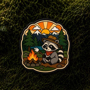 May include: A die-cut sticker featuring a cartoon raccoon wearing a brown hat and red bandana, sitting by a campfire while roasting a marshmallow on a stick. The background shows green pine trees, a blue lake, and snow-capped mountains under a sun.