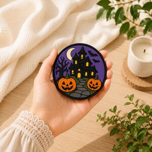 Może przedstawiać: Okrągła gumowa podstawka pod kubek z motywem Halloween, przedstawiająca czarny nawiedzony dom, dwie pomarańczowe dynie, szarą kamienną ścieżkę, bezlistne drzewo i dwa czarne nietoperze na tle fioletowego nieba z żółtym półksiężycem.