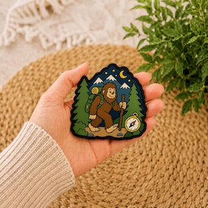 May include: A die-cut sticker featuring a brown Bigfoot character hiking through a forest with a green backpack and a wooden walking stick. The scene includes snow-capped mountains, a yellow crescent moon, stars, and a gold compass.